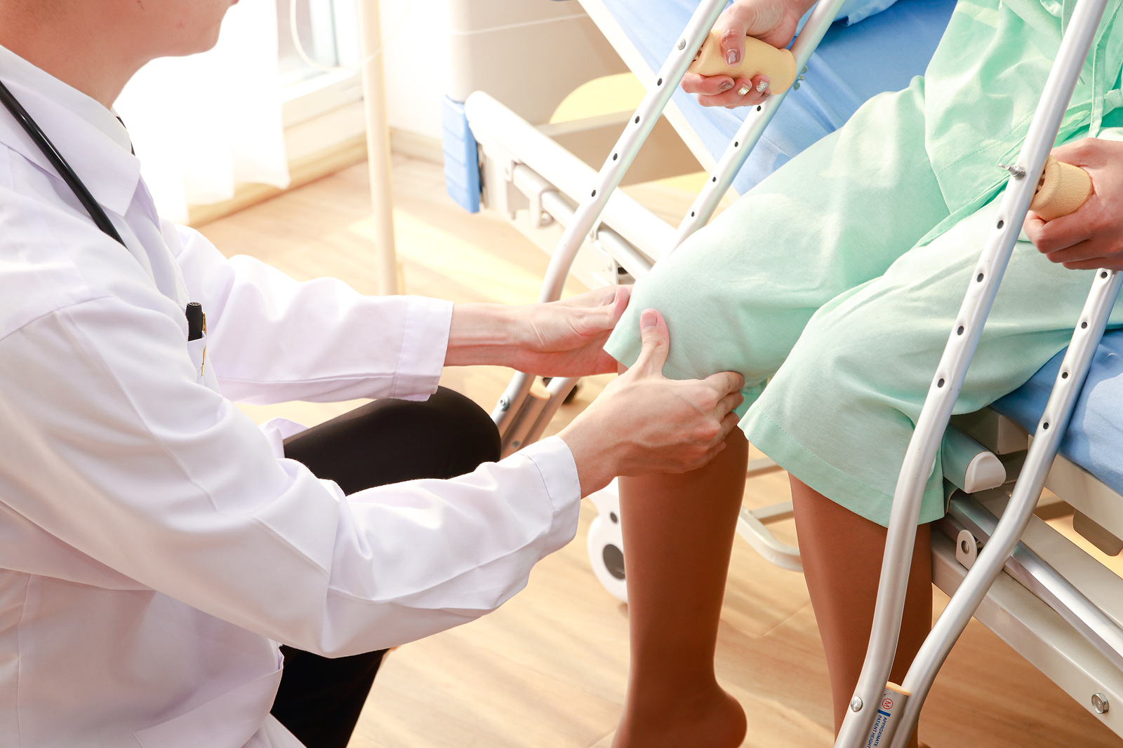 Doctor examining a female patient’s knee for pain and mobility issues associated with osteoarthritis while she sits on a nursing bed holding a cane.